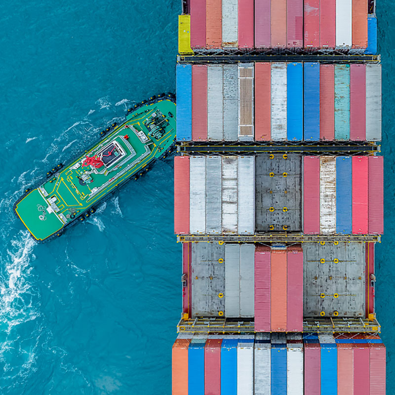 Aerial view of a tugboat maneuvering a massive cargo ship loaded with colorful shipping containers in blue water.