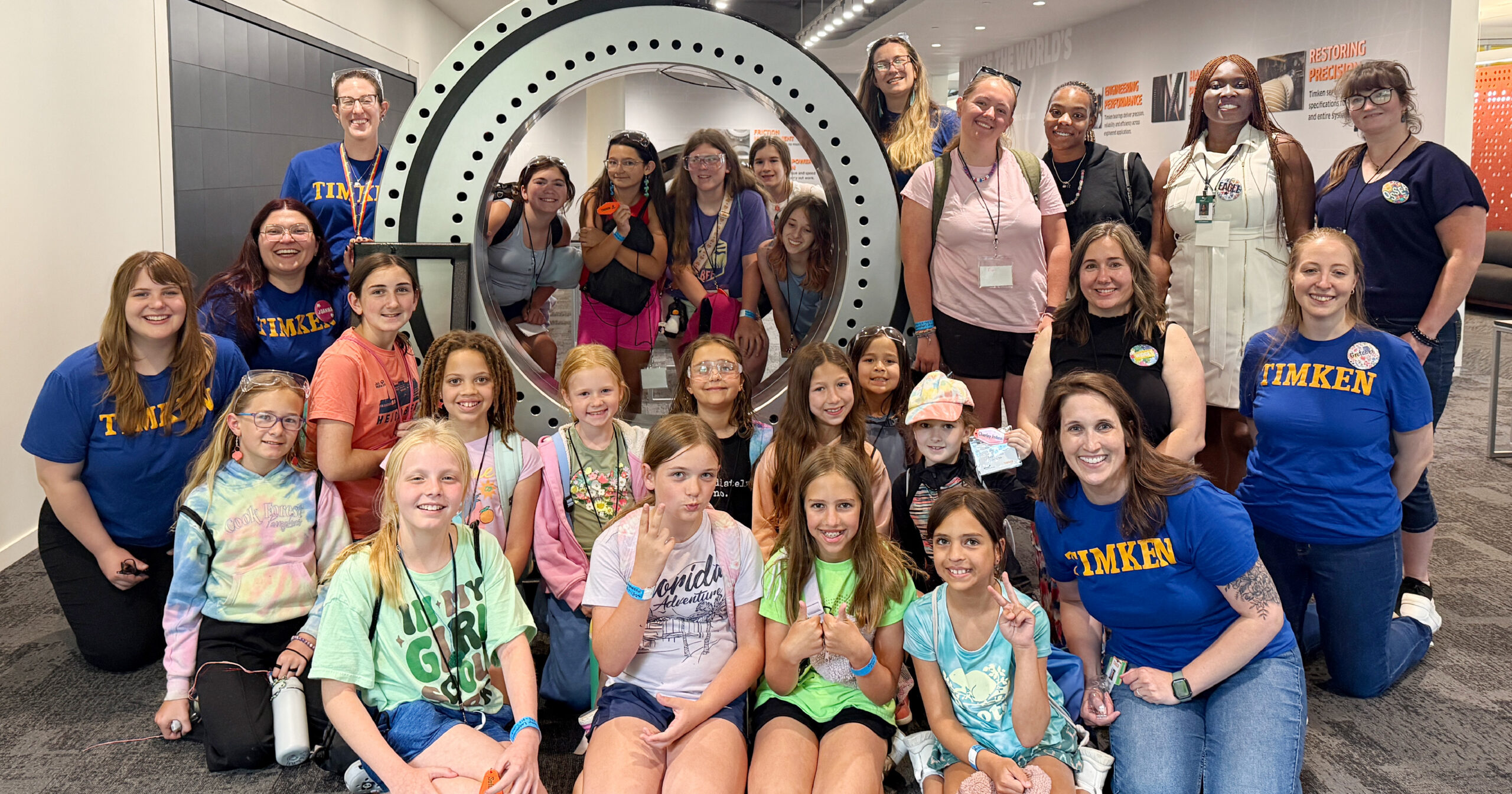 A group photo of girl scouts flanked by Timken volunteers and scout leaders, in front of a 6-foot diameter Timken bearing.